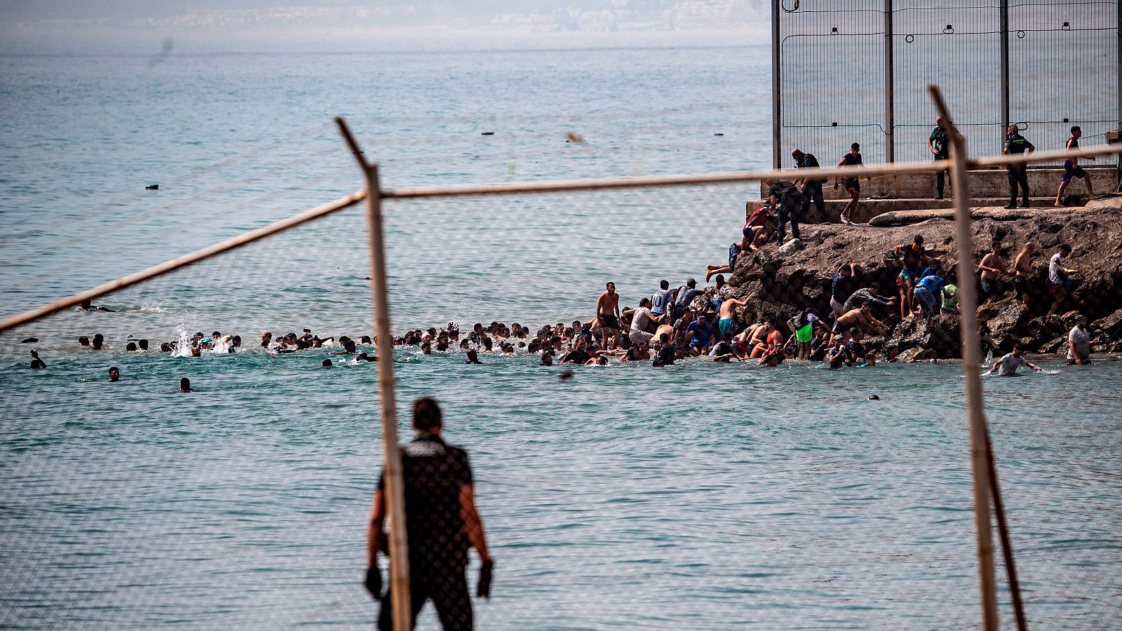 Cinco Continentes - El poder de Marruecos en torno al control migratorio Multitud cruza frontera marítima en Ceuta, España. Personas nadan y trepan rocas hacia una valla, mientras agentes observan.
