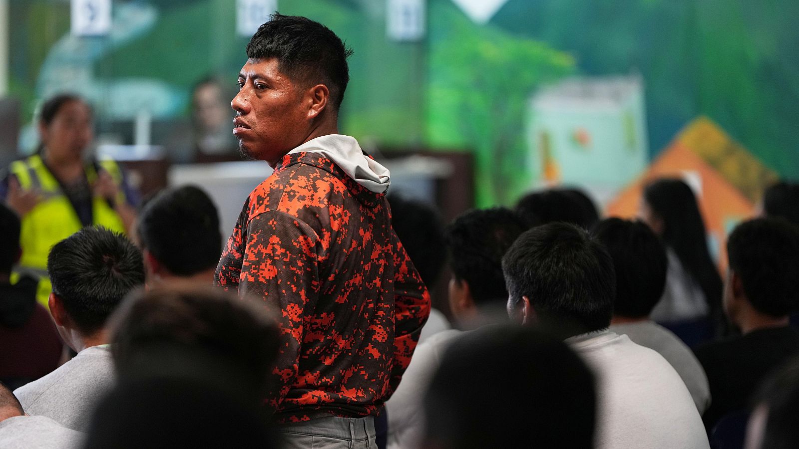 Hombres deportados esperan en un centro de procesamiento en Guatemala. Un hombre con sudadera de camuflaje es el foco principal, mientras una persona con chaleco amarillo dirige al grupo frente a un mural.