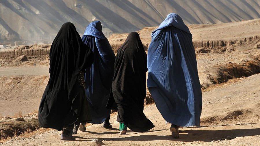 17218708 Cuatro mujeres con túnicas oscuras caminan por un camino de tierra en un paisaje árido y montañoso, con sombras alargadas.
